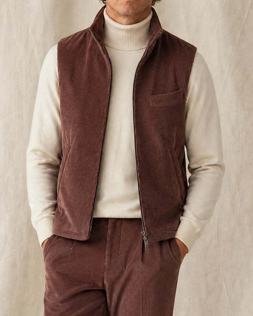 Mann trägt eine braune Bodywarmer Cordweste mit passender Cordhose, hellem Rollkragen und beigen Loafern vor neutralem Studiohintergrund