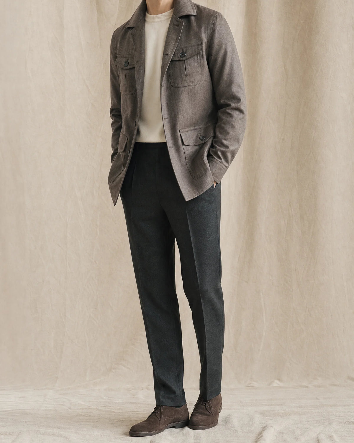 Mann trägt ein graues Overshirt mit hellem Strickpullover, dunkler Stoffhose und braunen Schuhen vor neutralem Studiohintergrund