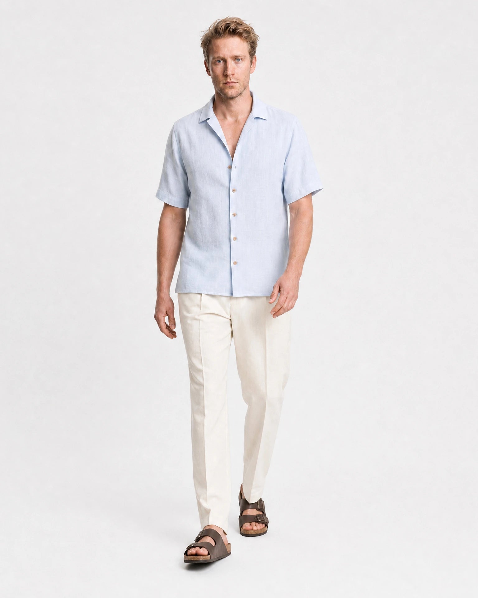 Hellblaues Leinen Kurzarmhemd Herren in Laufpose, cremefarbene Leinenhose und braune Sandalen, sommerlicher Look