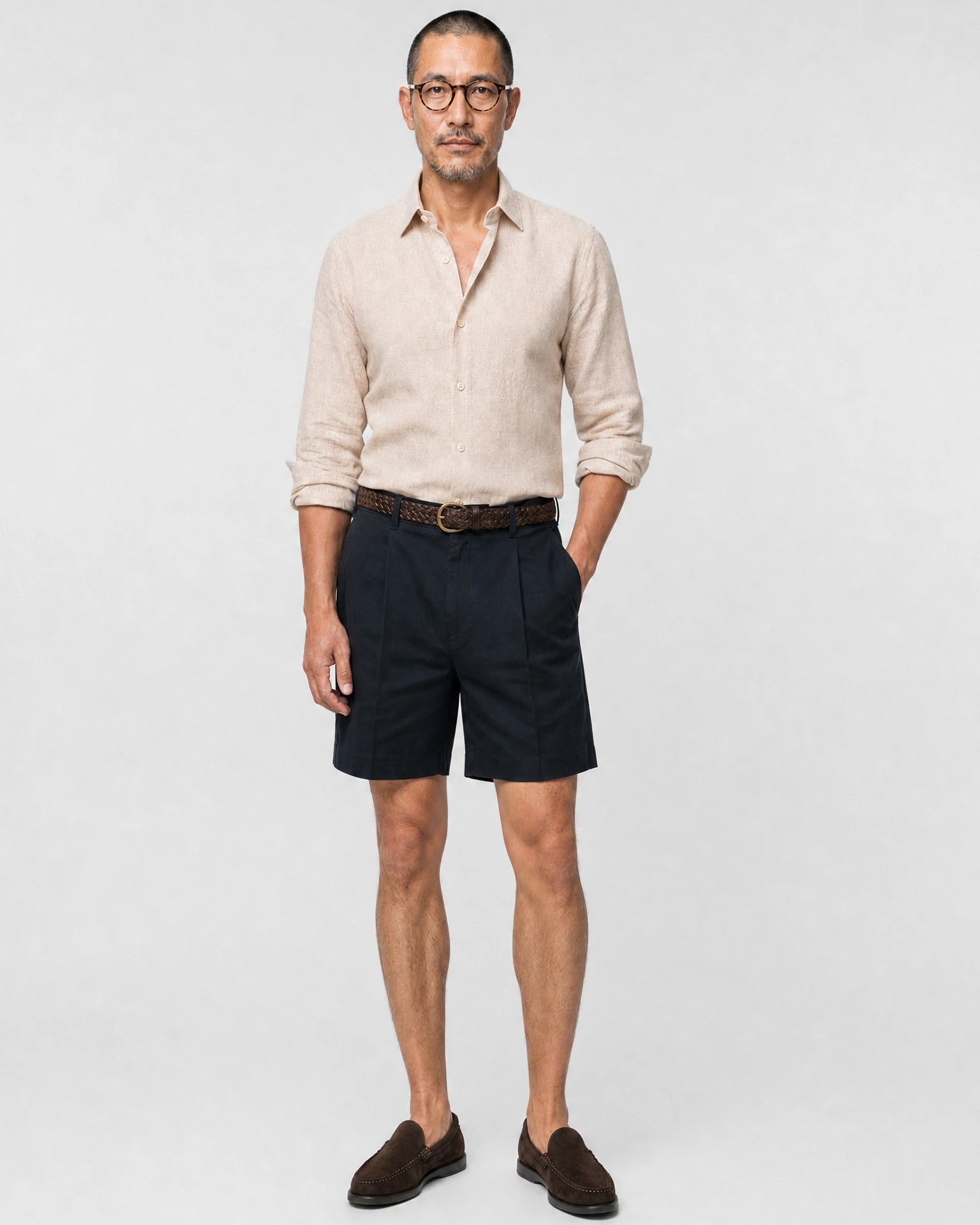 Sand-beiges Leinen Herringbone Hemd Herren Ganzkörper frontal, navy Bundfaltenshorts, brauner Flechtgürtel und braune Suede-Loafer