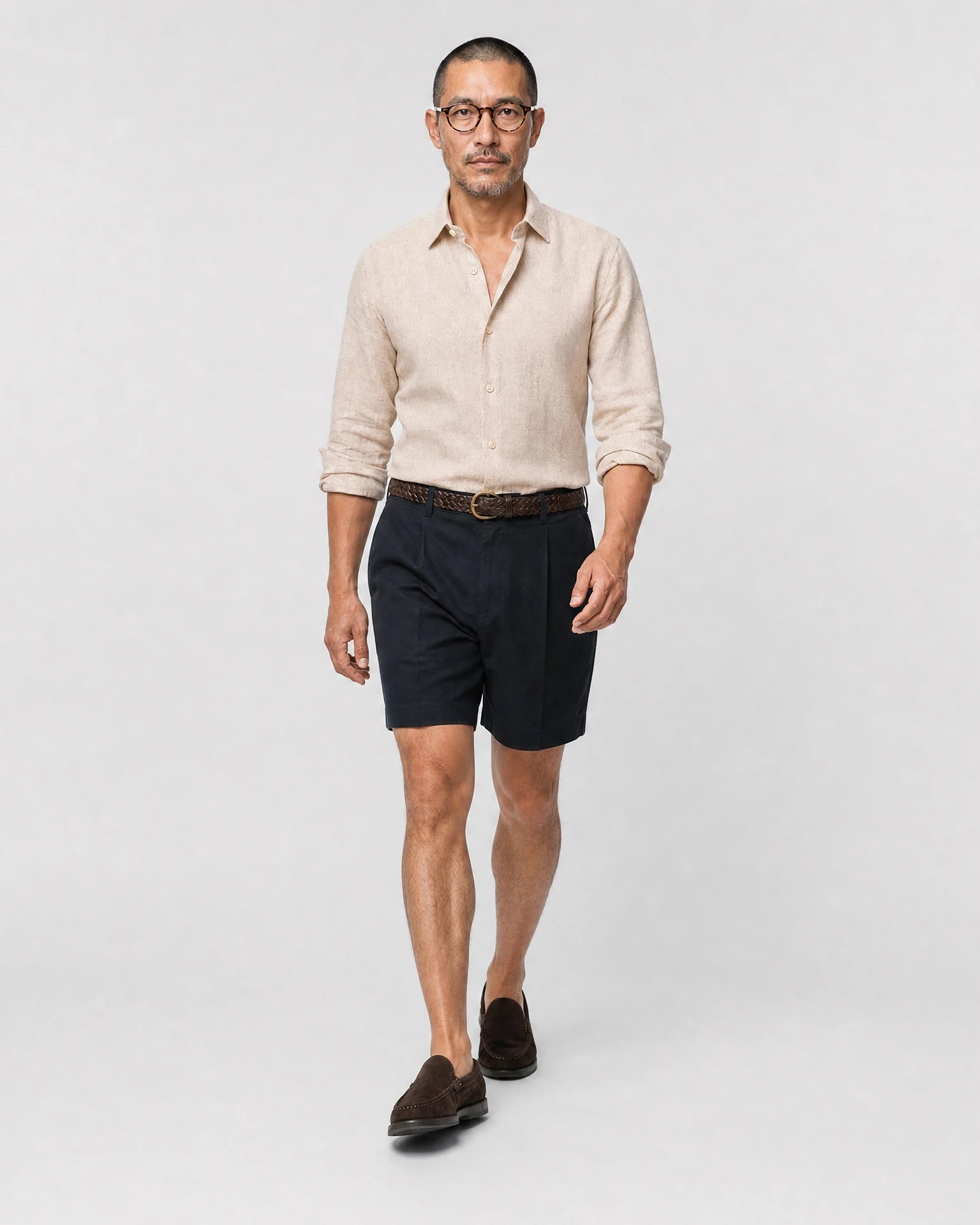 Sand-beiges Leinen Herringbone Hemd Herren in Laufpose, navy Bundfaltenshorts mit braunem Flechtgürtel und braunen Suede-Loafern