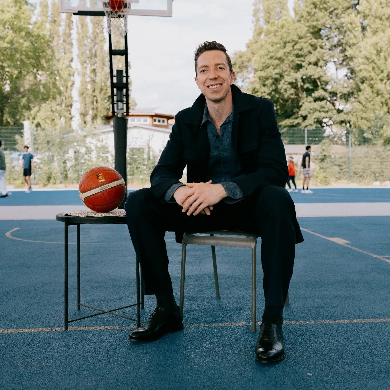 Mann im dunklen Maßanzug sitzt lächelnd auf einem Basketballplatz, neben ihm ein Basketball vor dem Korb