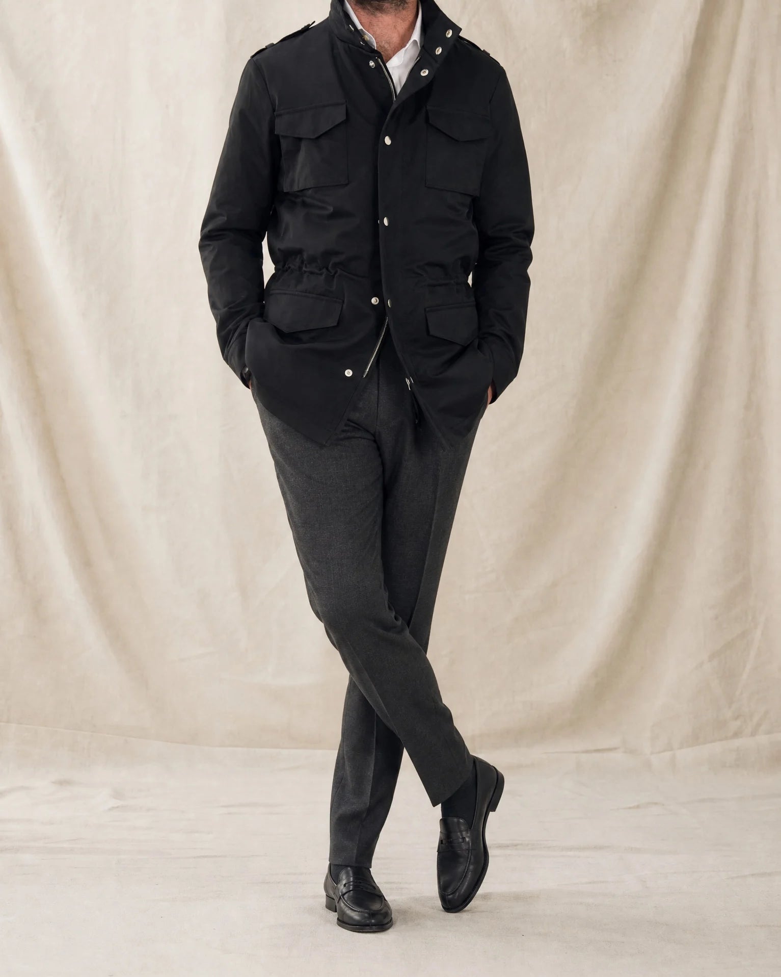 Mann trägt eine dunkle Field Jacket mit grauer Stoffhose, weißem Hemd und schwarzen Loafern vor neutralem Studiohintergrund