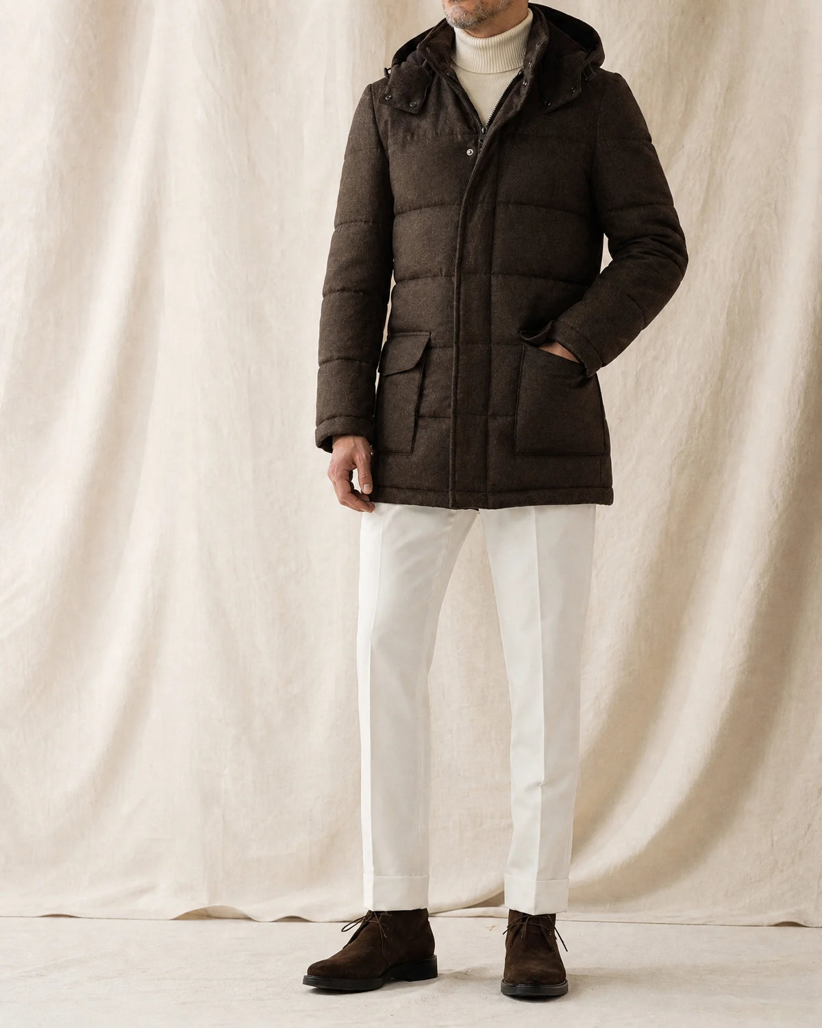 Mann trägt eine braune Steppjacke mit Kapuze, Rollkragen, weißer Stoffhose und braunen Boots vor neutralem Studiohintergrund