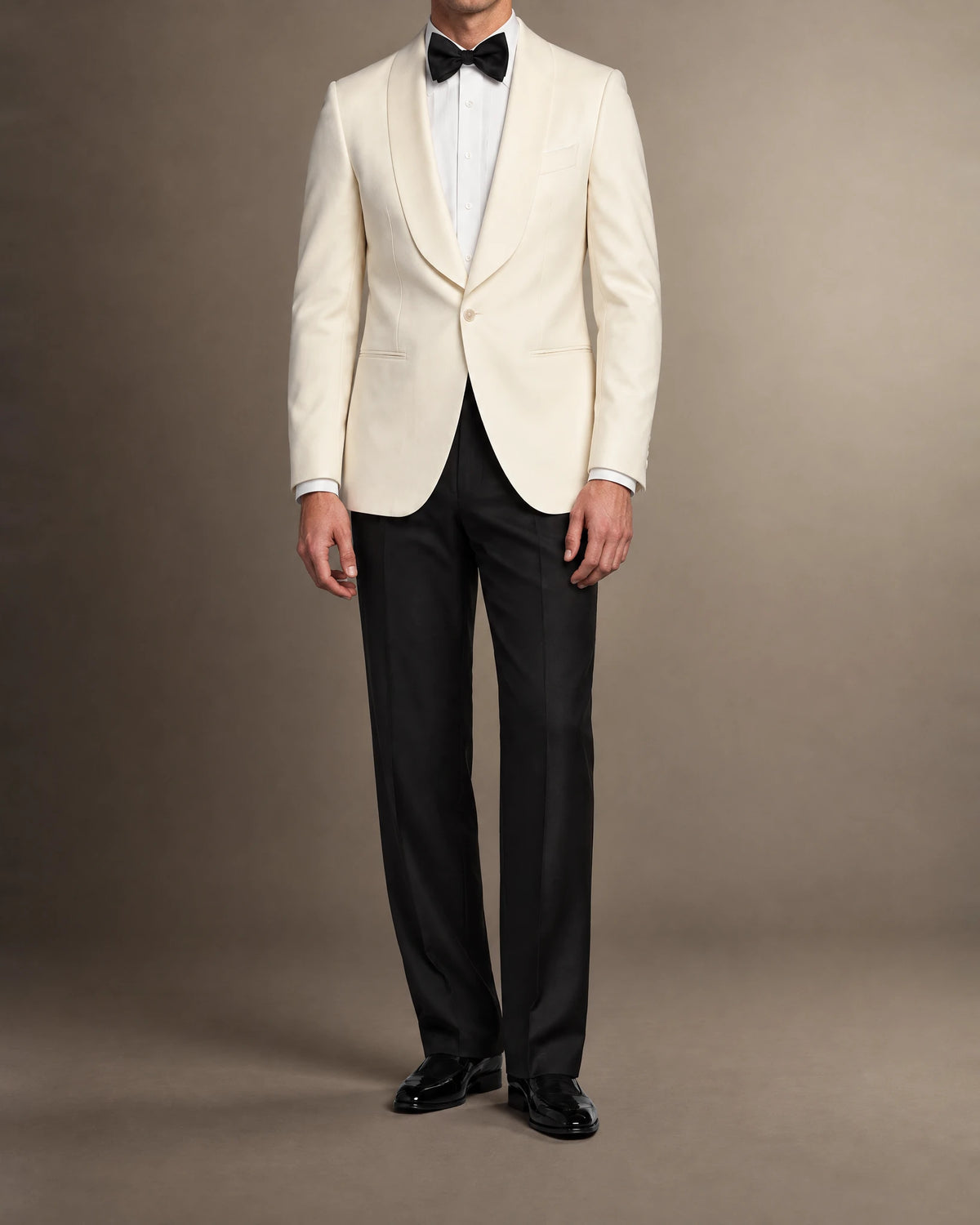 Mann im cremeweißen Dinnerjacket mit Schalkragen, schwarzer Smokinghose, Fliege und Lack-Loafern in frontaler Ganzkörperansicht
weisses-dinnerjacket-schalkragen-detail-fliege.webp