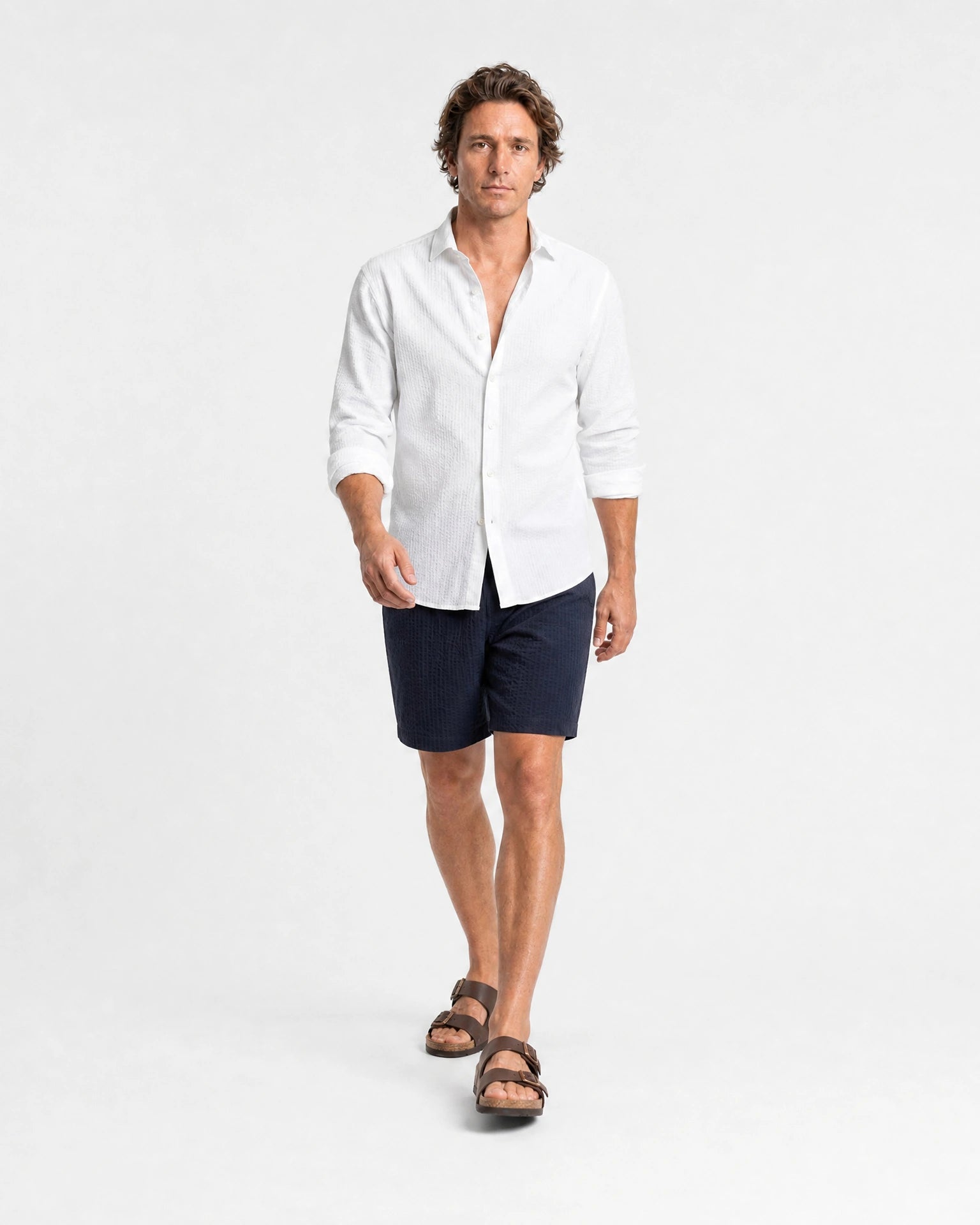 Weißes Seersucker Hemd Herren Ganzkörper in Laufpose, navy Seersucker-Shorts und braune Ledersandalen, sommerlicher Look