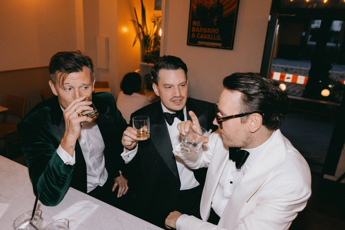 Drei Männer tragen elegante Smoking-Looks mit Fliege an einer Bar in warm beleuchteter Abendatmosphäre.