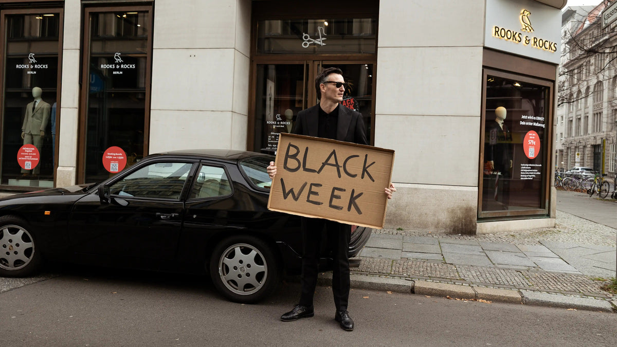 Black-Week-Aktion von ROOKS & ROCKS vor dem Store in Berlin Friedrichstraße mit Mann im schwarzen Anzug, Schild und schwarzem Porsche.