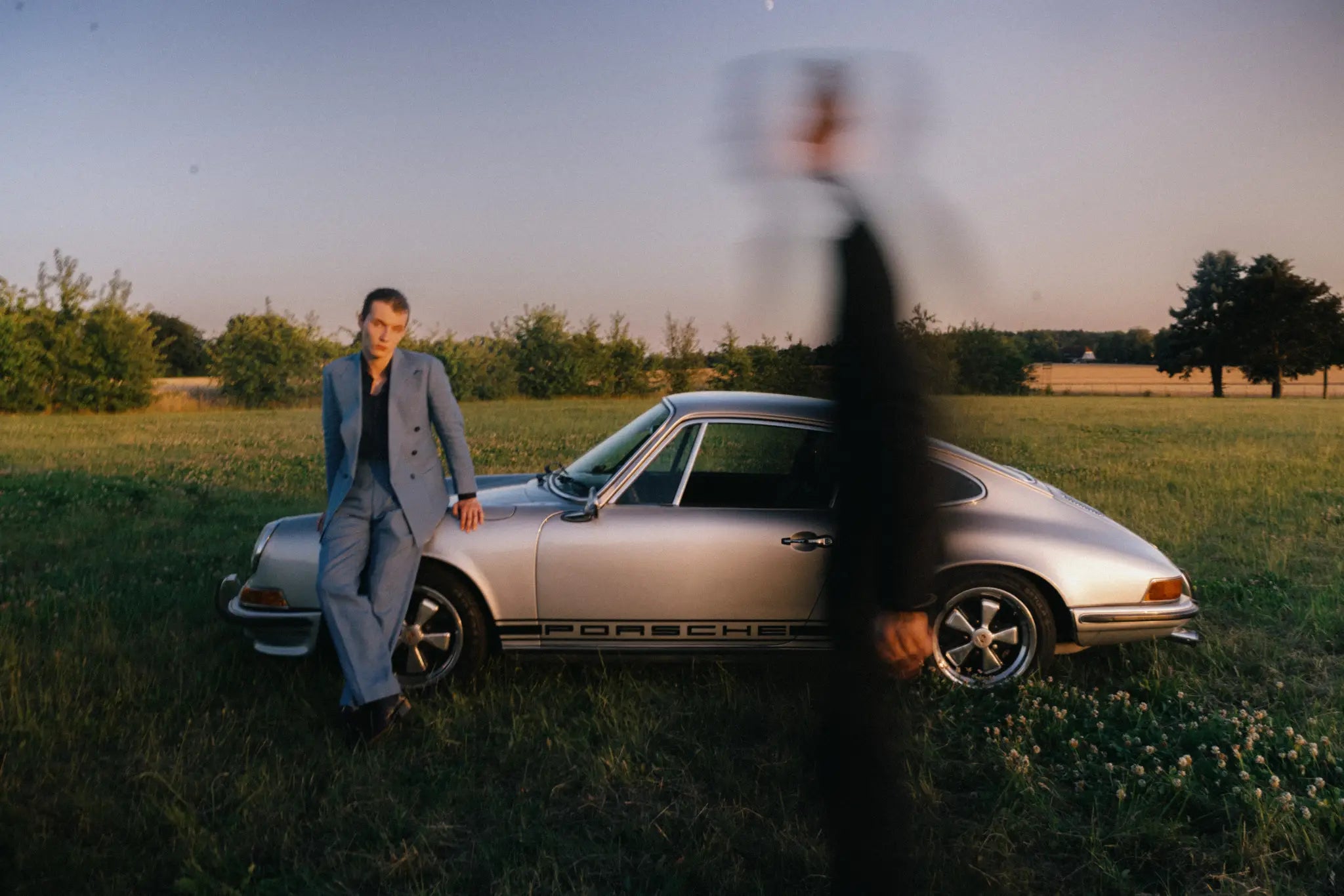 Sommerlicher Herren-Maßanzug in Hellblau vor klassischem Porsche 911 F-Serie in ländlicher Editorial-Kulisse bei warmem Abendlicht.