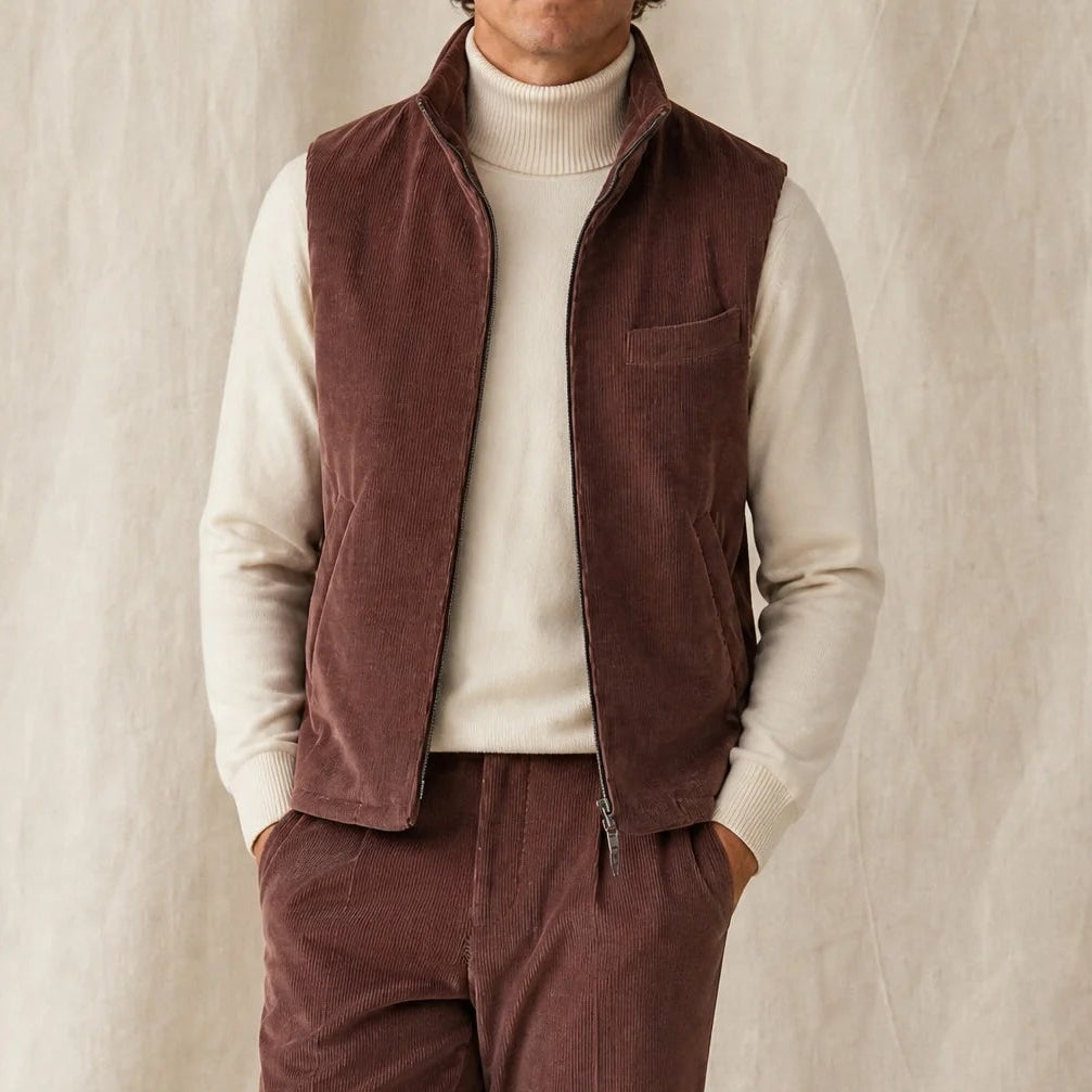 Mann trägt eine braune  Bodywarmer Cordweste mit passender Cordhose, hellem Rollkragen und beigen Loafern vor neutralem Studiohintergrund