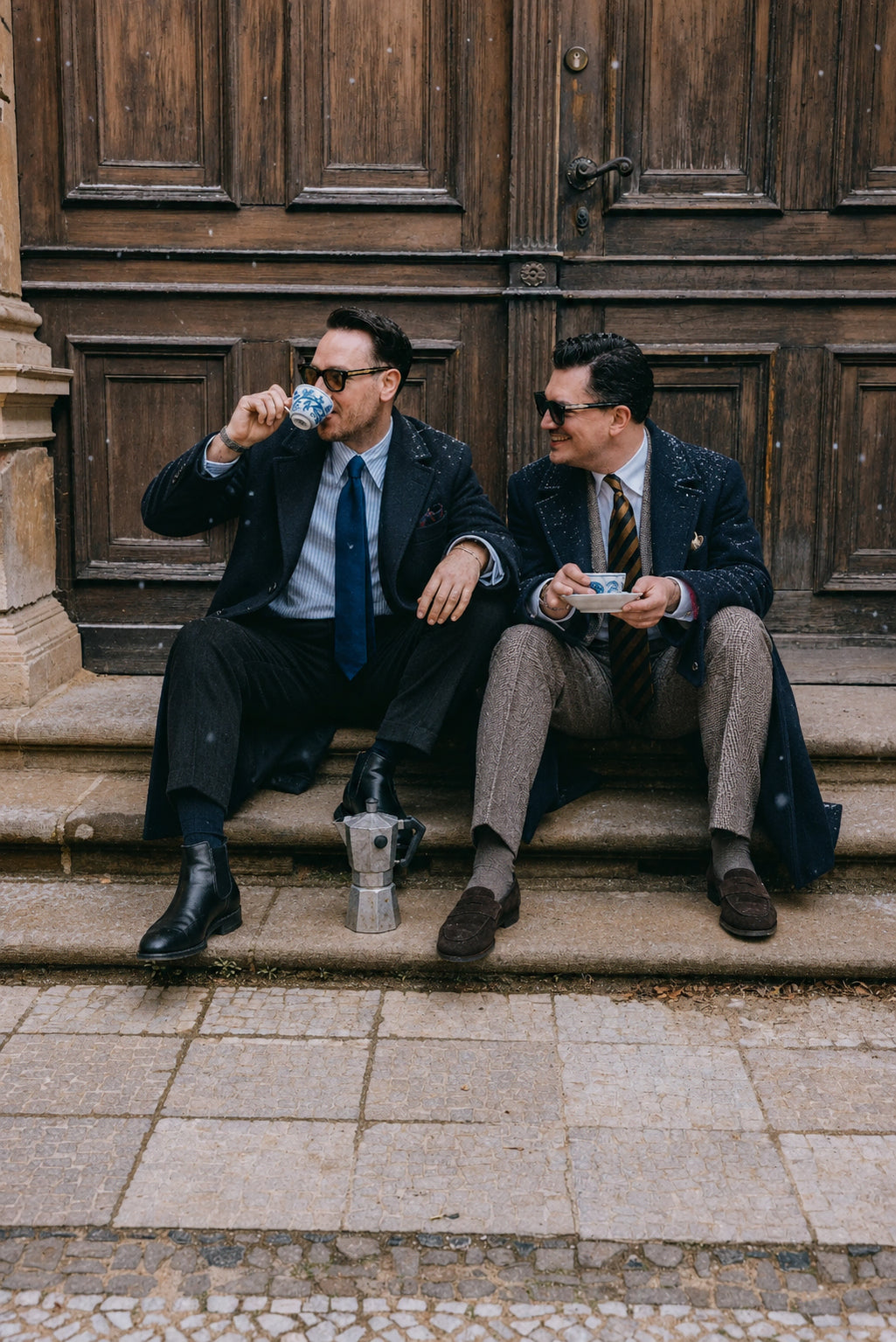 Zwei Männer tragen elegante Herrenlooks mit Mantel, Sakko, Krawatte und Loafern vor historischer Kulisse als stilvolle Winter-Inspiration.