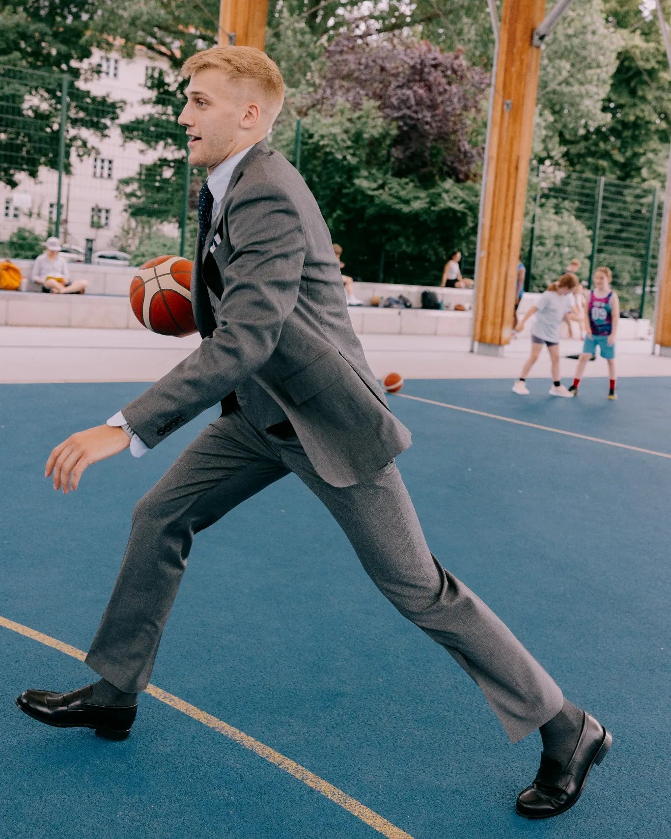 Mann im grauen Maßanzug dribbelt mit einem Basketball auf einem urbanen Court und kombiniert Business-Stil mit sportlicher Bewegung.