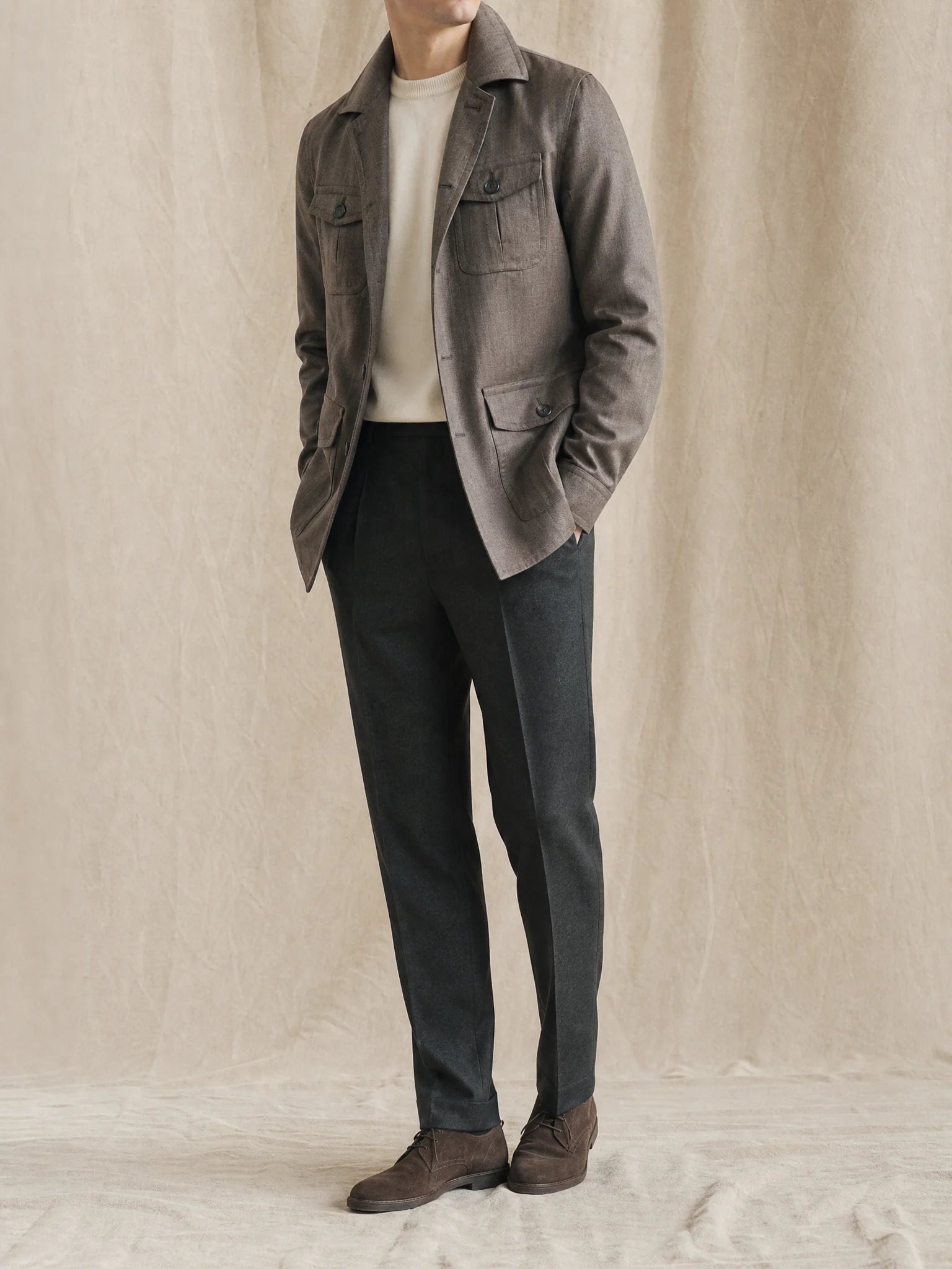 Mann trägt ein graues Overshirt mit hellem Strickpullover, dunkler Stoffhose und braunen Schuhen vor neutralem Studiohintergrund