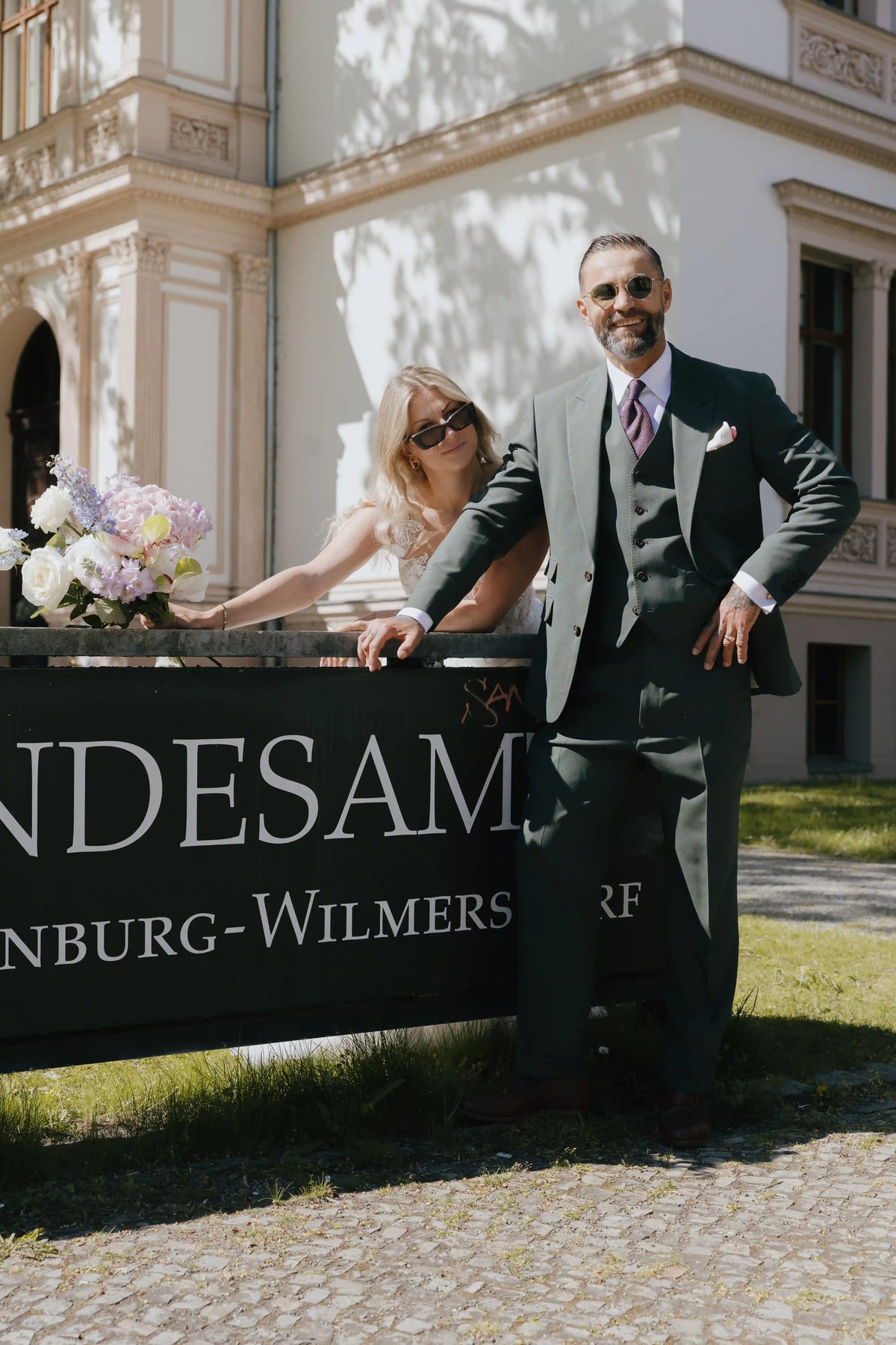 Brautpaar vor eleganter Schlosskulisse, der Bräutigam trägt einen grünen dreiteiligen Hochzeitsanzug mit Krawatte und Einstecktuch