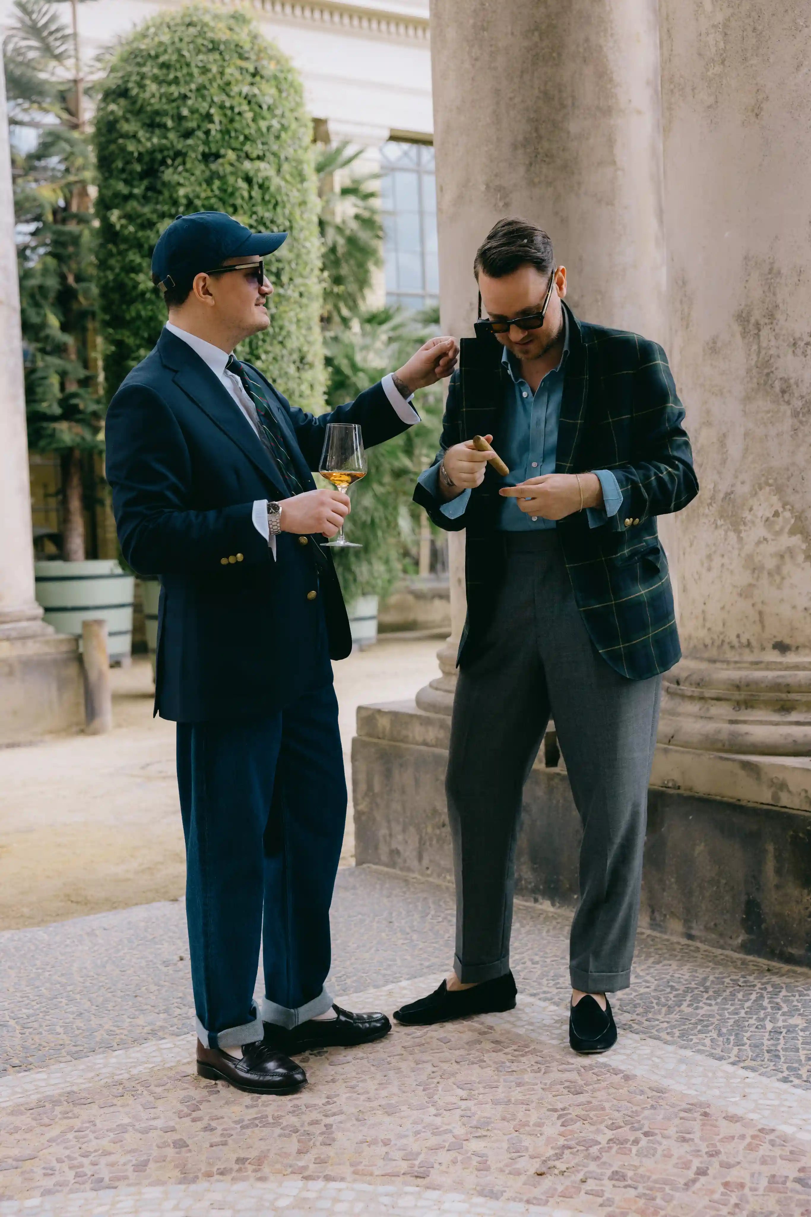Zwei Männer in klassischen ivy league Sommerlooks genießen einen stilvollen Moment mit Glas und Zigarre vor historischen Säulen