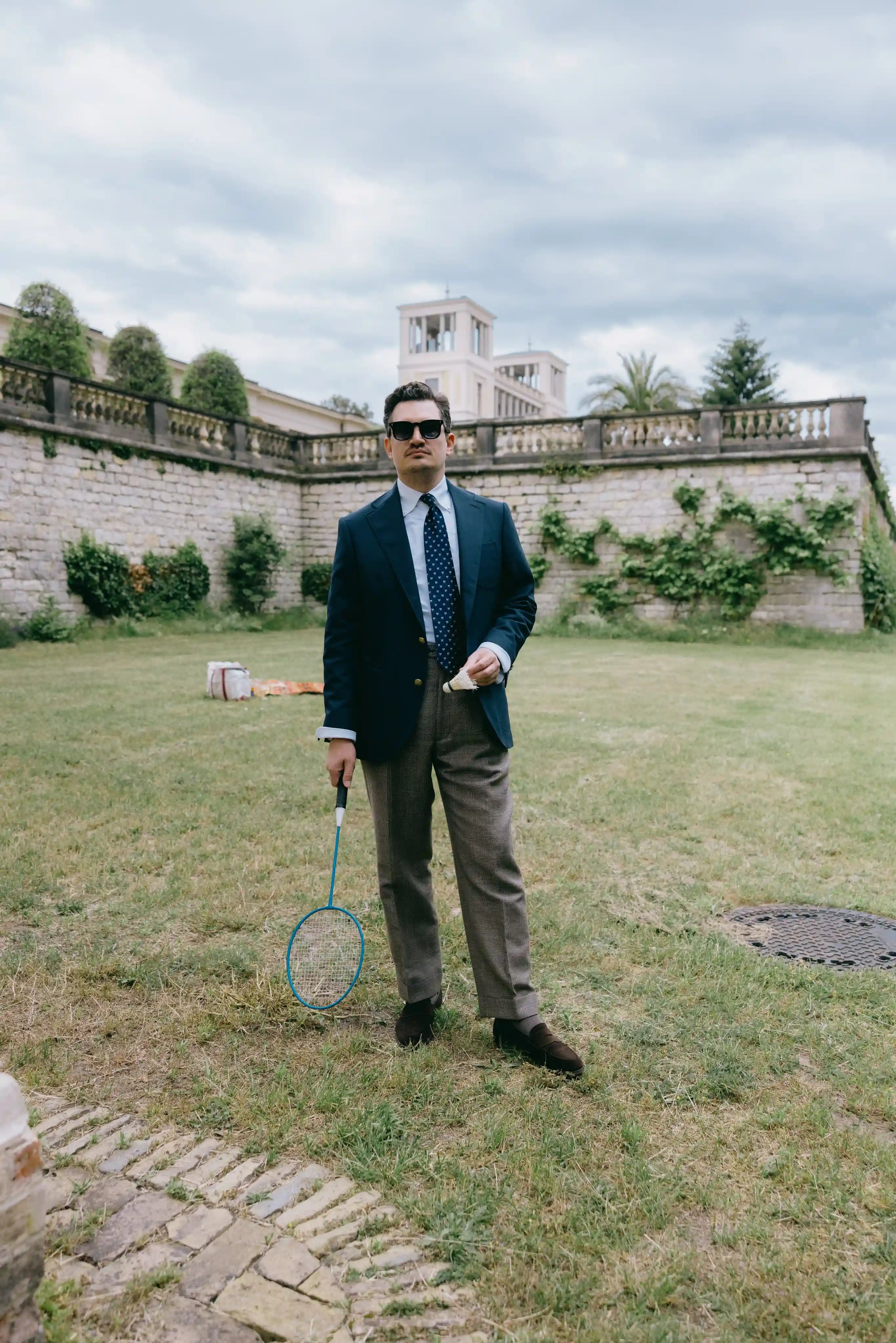 Stilvoller Herrenlook mit navyblauem Blazer, gemusterter Krawatte und Badmintonschläger in sommerlicher Parkkulisse