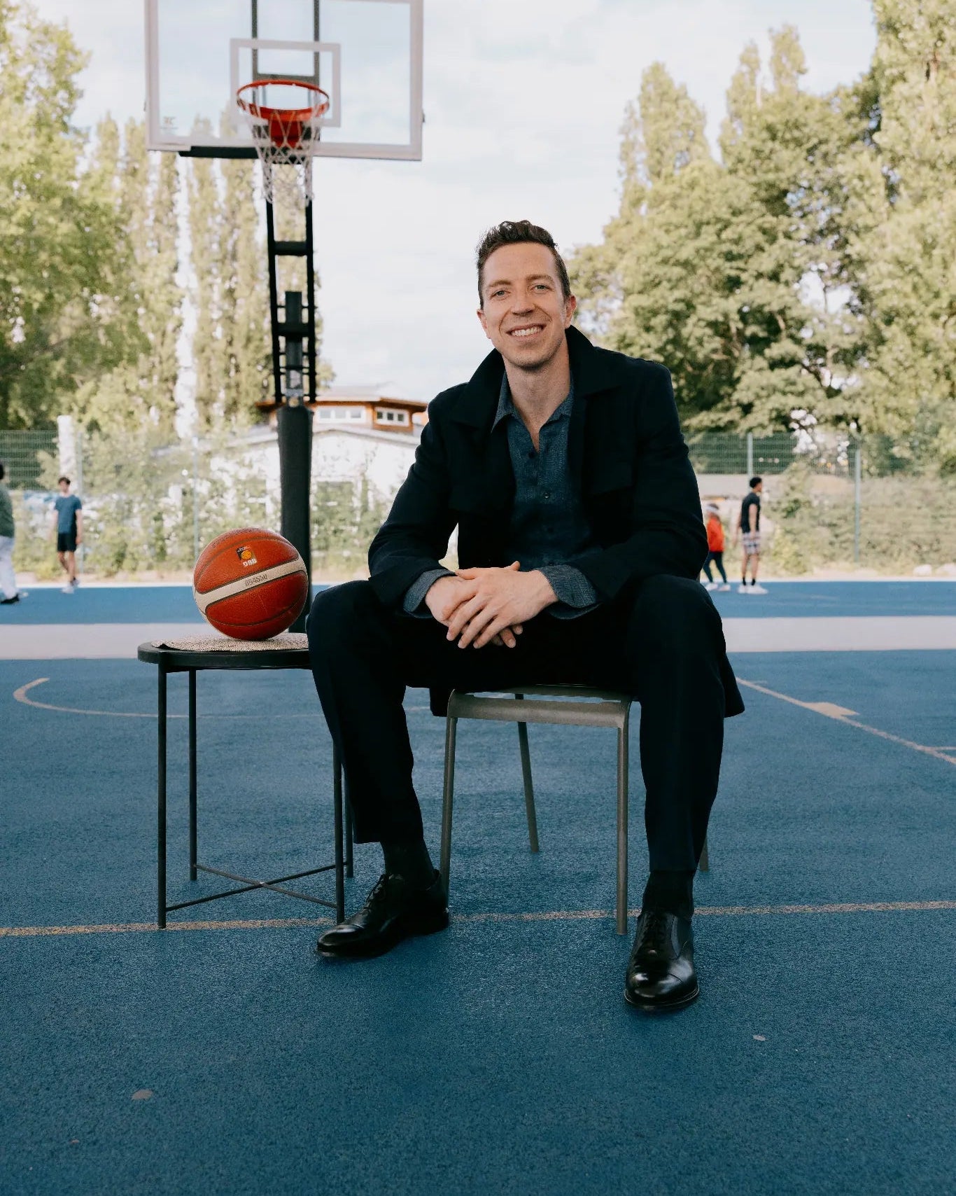 Mann im dunklen Maßanzug sitzt lächelnd auf einem Basketballplatz, neben ihm ein Basketball vor dem Korb