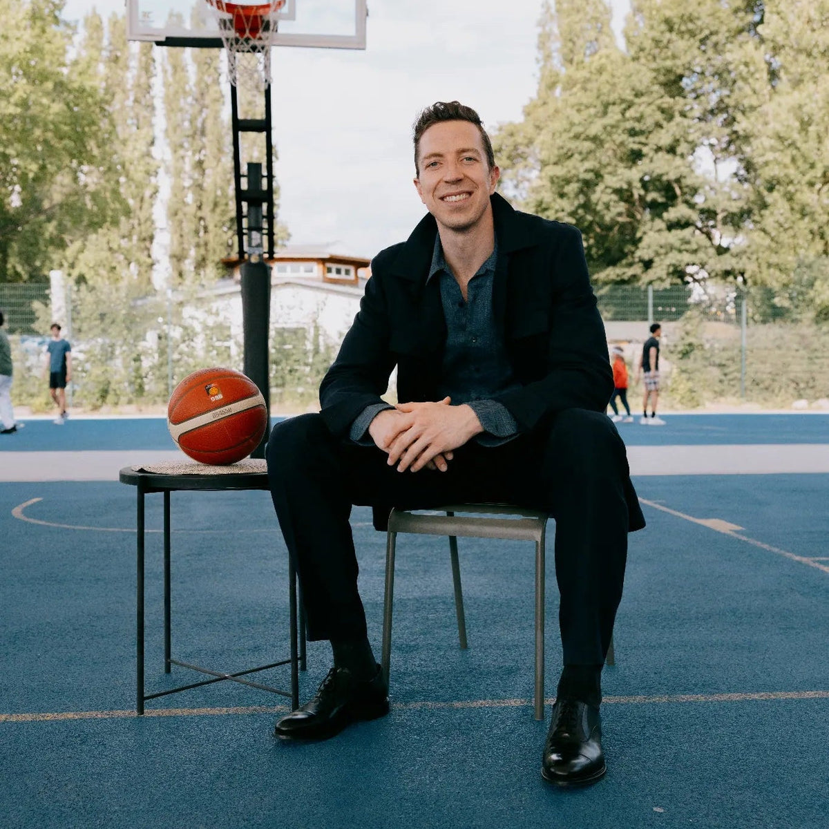 Mann sitzt lächelnd auf Stuhl auf einem Basketballfeld, neben ihm ein Basketball