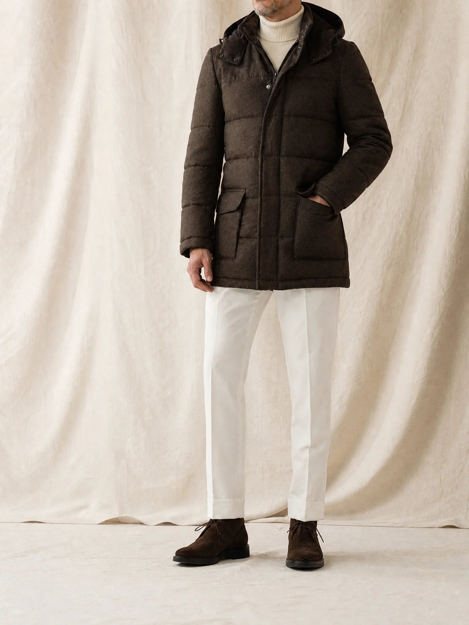 Mann trägt eine braune Steppjacke mit Kapuze, Rollkragen, weißer Stoffhose und braunen Boots vor neutralem Studiohintergrund