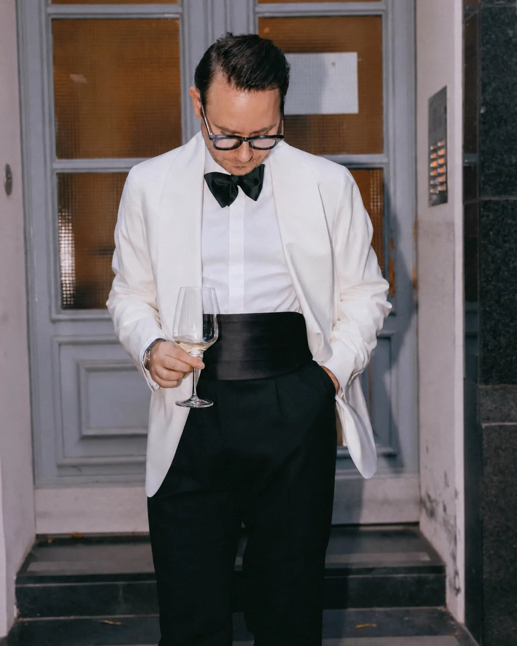 Massgeschneidertes weißes Dinner Jacket für Herren mit schwarzer Fliege, Kummerbund, Smokinghose und Weinglas als eleganter Black-Tie-Abendlook.