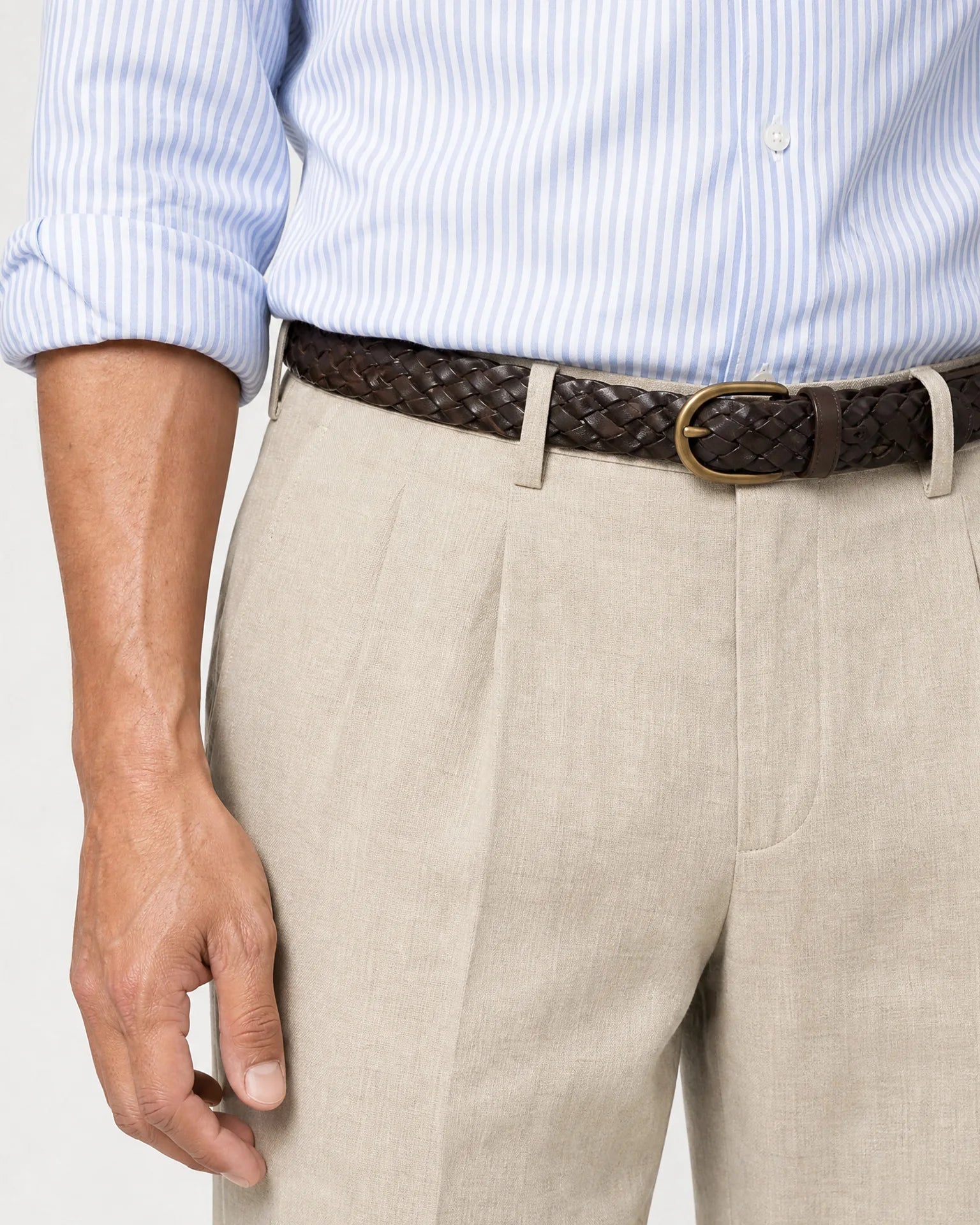 Detailaufnahme der beigen Leinenhose mit Bundfalten, geflochtenem dunkelbraunem Gürtel und hellblau gestreiftem Hemd.