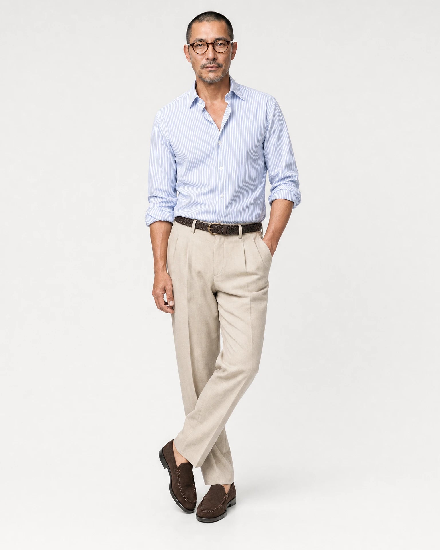 Ganzkörperansicht eines Models in beiger Leinenhose mit hellblau gestreiftem Hemd, geflochtenem dunkelbraunem Gürtel und braunen Loafern.