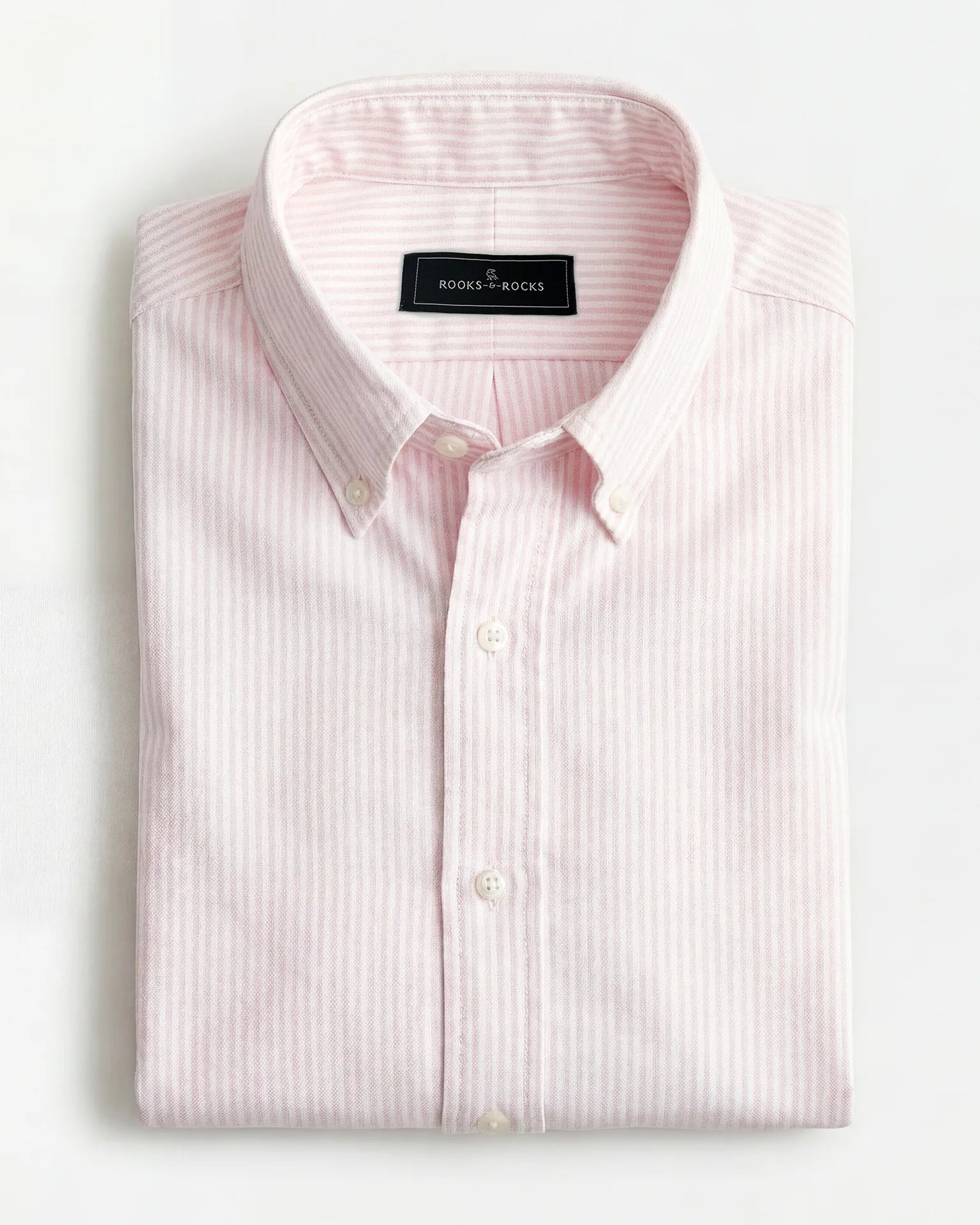 Gefaltetes Rooks & Rocks Oxford-Hemd in Rosa-Weiß gestreift mit Button-Down-Kragen auf hellem Hintergrund.