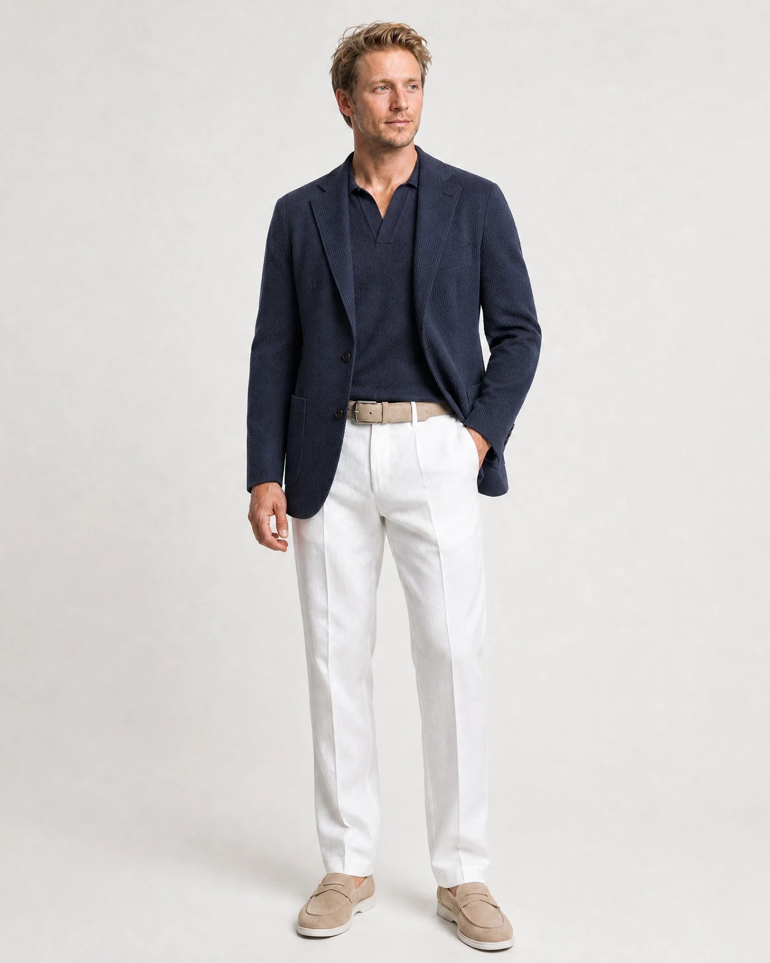 Weiße Leinenhose kombiniert mit dunkelblauem Sakko, navy Poloshirt, beigem Gürtel und hellen Loafern.