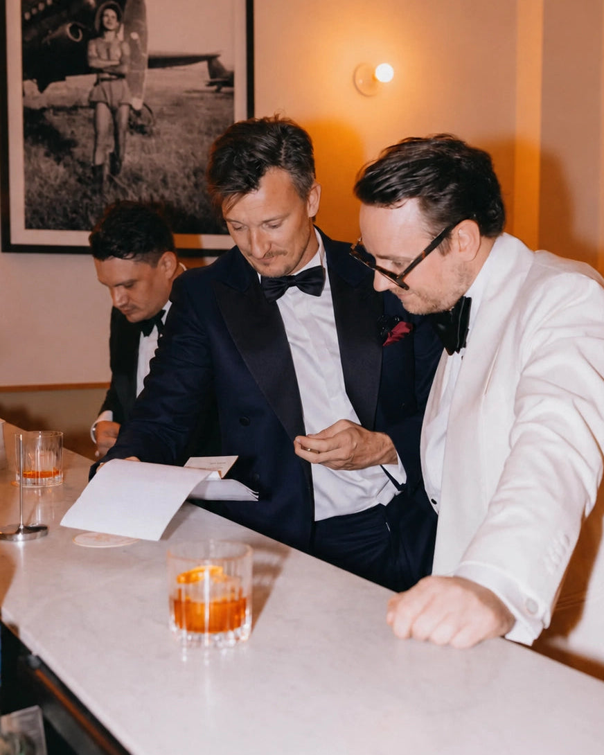 Elegante Black-Tie-Inspiration für Herren mit schwarzem Smoking, weißem Dinner Jacket, Fliege und Drinks an einer stilvollen Bar.