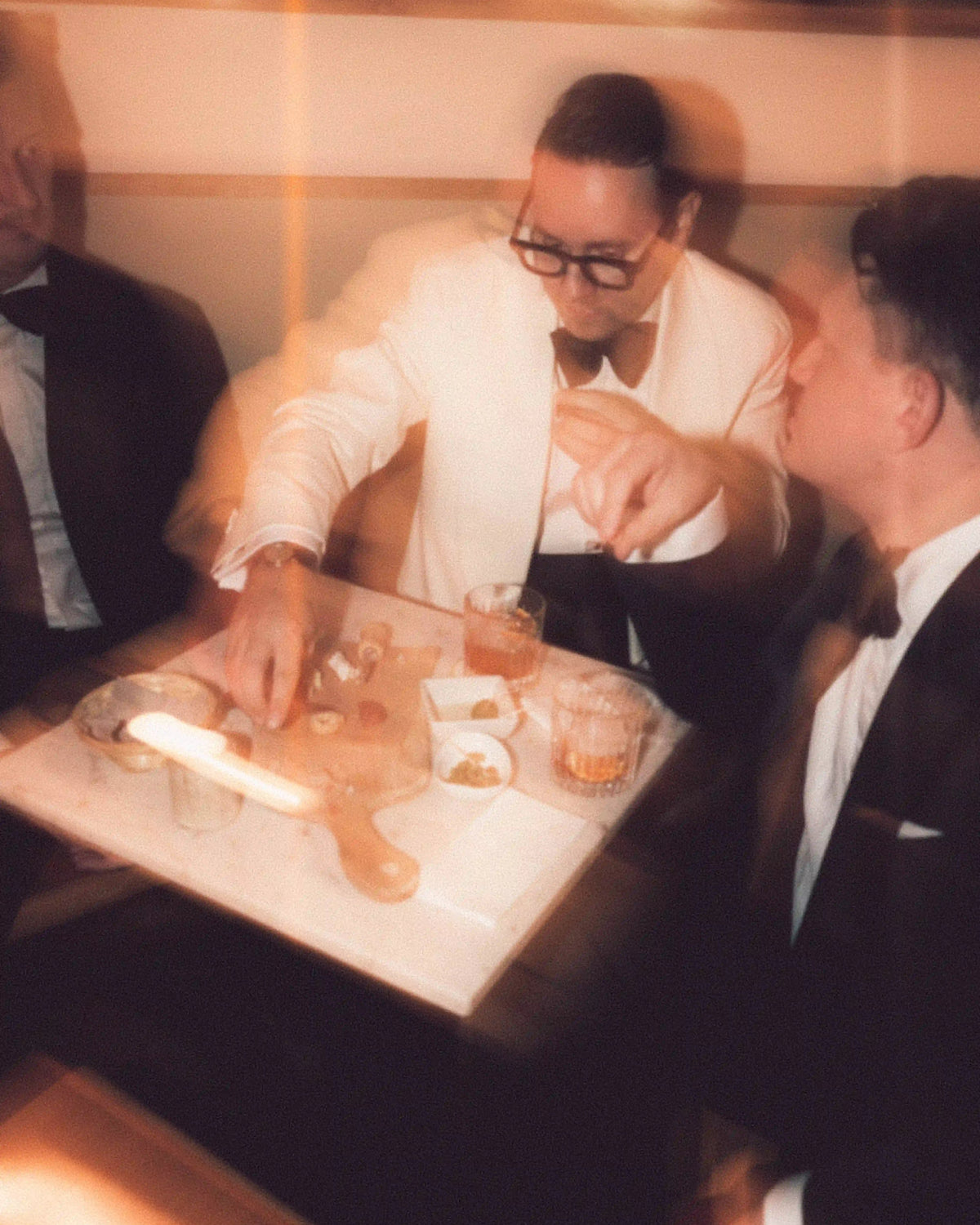 Drei Männer in eleganten Smoking-Looks sitzen bei Drinks an einer Bar in warmer, unscharfer Candlelight-Atmosphäre.