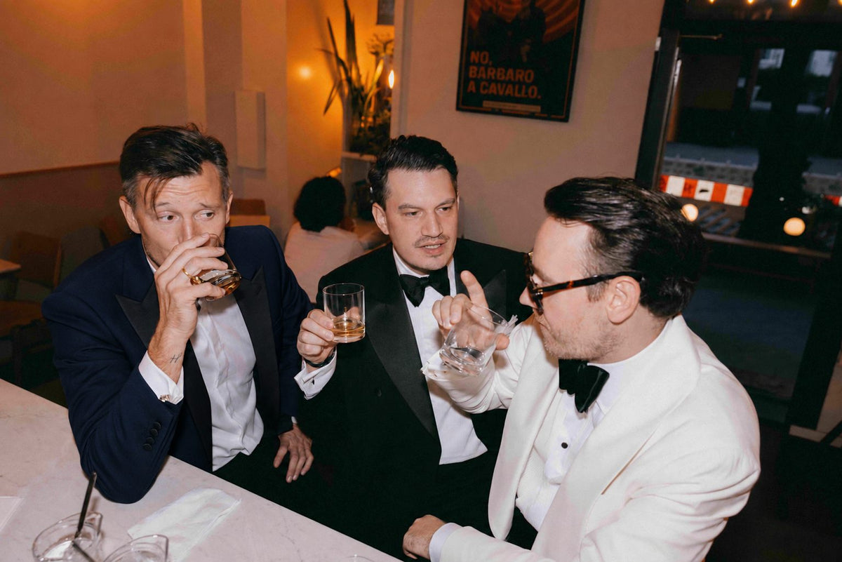 Elegante Black-Tie-Inspiration für Herren mit schwarzem Smoking, weißem Dinner Jacket, Fliege und Drinks an einer stilvollen Bar.