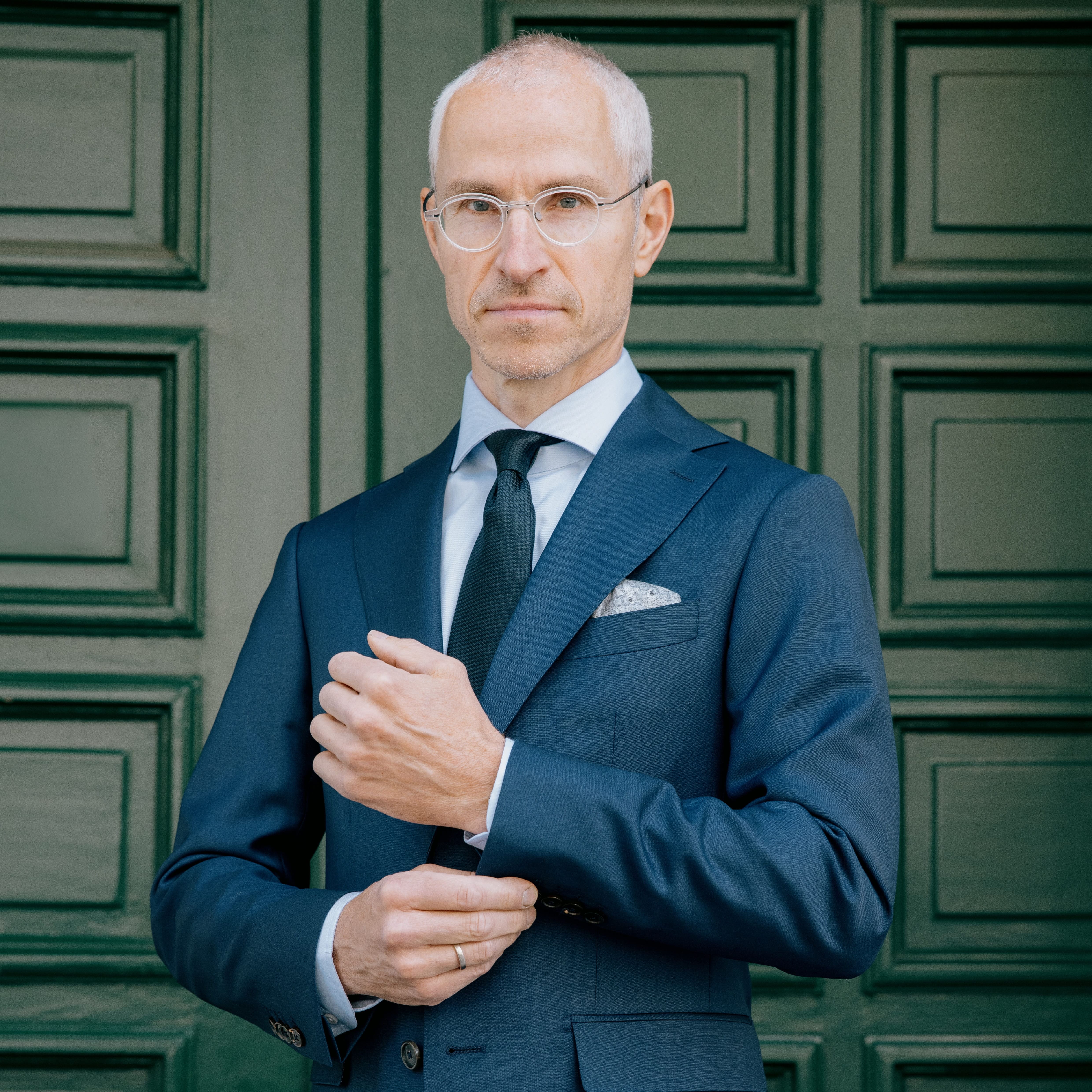 Älterer Herr mit grauen Haaren und Brille im blauen Anzug steht vor einer grünen Tür